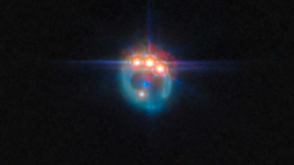 A small image of a galaxy distorted by gravitational lensing into a dim ring. At the top of the ring are three very bright spots with diffraction spikes coming off them, right next to each other: these are copies of a single quasar in the lensed galaxy, duplicated by the gravitational lens. In the centre of the ring, the elliptical galaxy doing the lensing appears as a small blue dot. The background is black and empty.